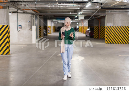 A woman wearing a hijab walks through a parking garage while looking at her phone and holding a coffee cup 135813728