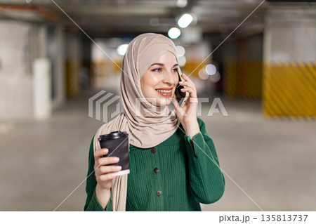 A smiling woman wearing a hijab is talking on her phone while holding a coffee cup in a parking garage A smiling woman wearing a hijab is talking on her phone while holding a coffee cup in a parking garage 135813737