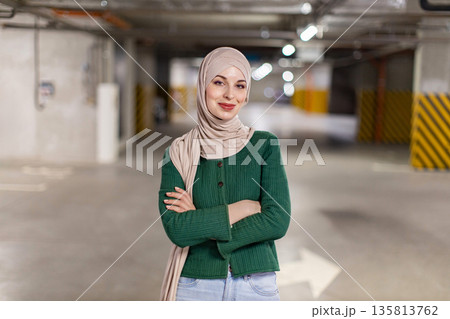A smiling woman wearing a hijab stands confidently in a parking garage with her arms crossed 135813762