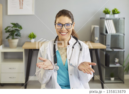 Smiling female doctor with stethoscope gesturing open hands in clinic office 135813870