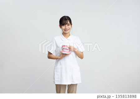 白背景の前で歯の模型を持って微笑む白衣を着た若い女性 白背景の前で歯の模型を持って微笑む白衣を着た若い女性 135813898