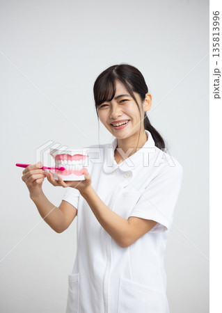 白背景の前で歯の模型と歯ブラシを持って微笑む白衣姿の女性 白背景の前で歯の模型と歯ブラシを持って微笑む白衣姿の女性 135813996