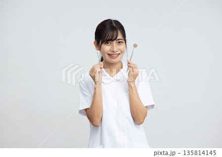 白背景の前で歯科用の器具を持って微笑む白衣を着た若い女性 135814145