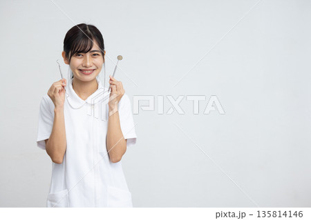 白背景の前で歯科用の器具を持って微笑む白衣を着た若い女性 白背景の前で歯科用の器具を持って微笑む白衣を着た若い女性 135814146