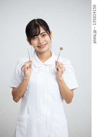 白背景の前で歯科用の器具を持って微笑む白衣を着た若い女性 白背景の前で歯科用の器具を持って微笑む白衣を着た若い女性 135814150