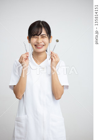 白背景の前で歯科用の器具を持って微笑む白衣を着た若い女性 135814151