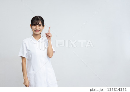 白背景の前で人差し指を立てて微笑む白衣を着た若い女性 135814351