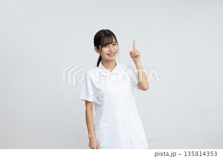 白背景の前で人差し指を立てて微笑む白衣を着た若い女性 白背景の前で人差し指を立てて微笑む白衣を着た若い女性 135814353