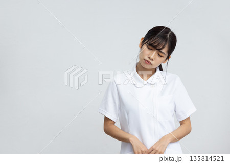 白背景の前で困った表情で首を傾げる白衣を着た若い女性 135814521