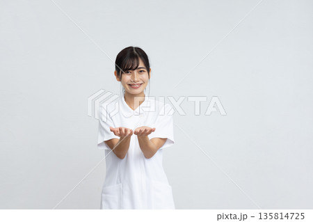 白背景の前で手をかざして微笑む白衣を着た若い女性 白背景の前で手をかざして微笑む白衣を着た若い女性 135814725