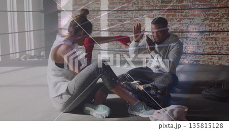 Trainee punching with red hand wraps and trainer holding pads in boxing ring, with bag, ropes Trainee punching with red hand wraps and trainer holding pads in boxing ring, with bag, ropes 135815128