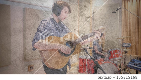 Strumming guitarist playing acoustic guitar in soundproof studio, with drummer behind red drum kit Strumming guitarist playing acoustic guitar in soundproof studio, with drummer behind red drum kit 135815169
