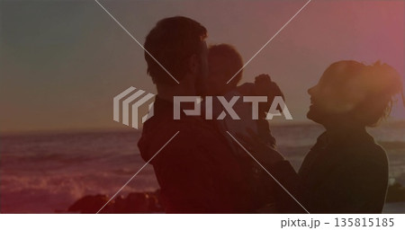 Parents holding baby in light onesie while standing at beach edge during orange sunset, silhouette 135815185