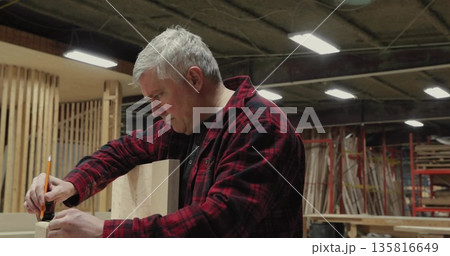 Carpenter wearing flannel marking wooden plank at workbench in wood shop, with pencil and square 135816649