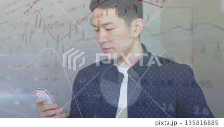 Holding smartphone, man wearing navy shirt scanning candlestick charts and line graphs in office 135816885