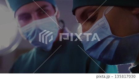 Leaning female surgeon wearing green scrubs masks caps under lamp in OR, with colleague blurred Leaning female surgeon wearing green scrubs masks caps under lamp in OR, with colleague blurred 135817030