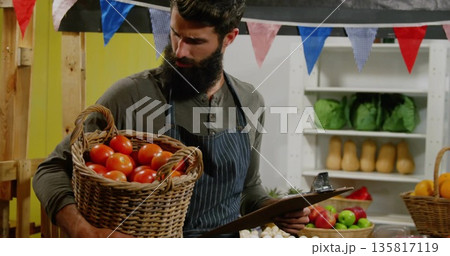 Bearded vendor standing and holding basket of tomatoes and clipboard in market stall, with produce Bearded vendor standing and holding basket of tomatoes and clipboard in market stall, with produce 135817119