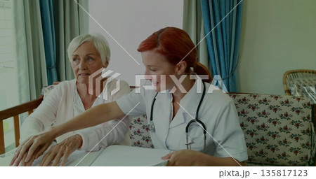 Pointing healthcare worker guiding patient on floral sofa reviewing documents, with stethoscope 135817123