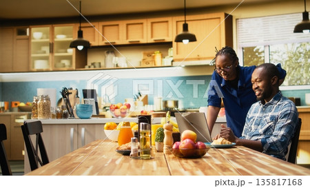 African American freelancer sitting at a kitchen table with a laptop, sending emails and explaining business tasks to his girlfriend. Couple enjoys a relaxed home lifestyle with coffee. African American freelancer sitting at a kitchen table with a laptop, sending emails and explaining business tasks to his girlfriend. Couple enjoys a relaxed home lifestyle with coffee. 135817168