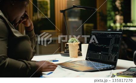 Black woman distressed about stock market trends going down and losing money, feeling scared and concerned about risk and bankruptcy. Studying forex data and being worried. Camera B. 135817270