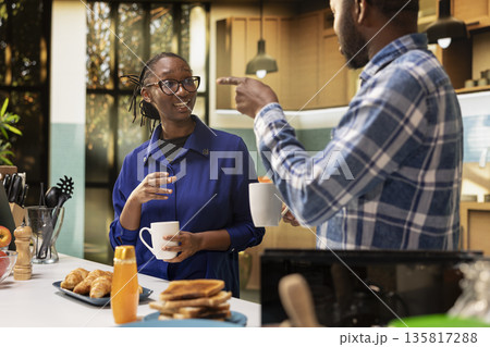 Black partners chatting and laughing over fresh morning coffee cups, preparing breakfast at the kitchen counter. Couple enjoying toast and homemade croissants with honey, morning routine. Black partners chatting and laughing over fresh morning coffee cups, preparing breakfast at the kitchen counter. Couple enjoying toast and homemade croissants with honey, morning routine. 135817288