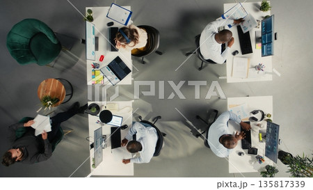 Top down view of staff in multinational company office reviews diagrams and business charts. Coworkers sharing insights and projections to enhance corporate planning. 135817339