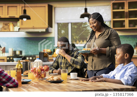 Smiling mother serving her kids and husband with bacon and eggs, family gathered around the kitchen table for breakfast enjoying meal under morning light. Peaceful routine with love. 135817366