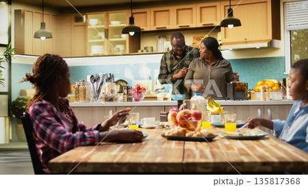Parents and children at home serving breakfast in the kitchen, mother preparing the meal at the kitchen counter. Family moment captures warmth, morning joy and being together. Camera B. 135817368