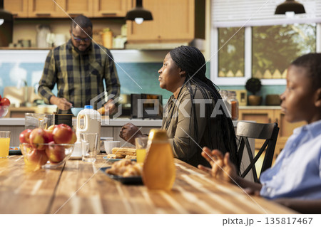 Family enjoying a weekend breakfast together in a sunny kitchen. Parents serve coffee, croissants and orange juice while children laugh, moment of love and happiness around the table. Family enjoying a weekend breakfast together in a sunny kitchen. Parents serve coffee, croissants and orange juice while children laugh, moment of love and happiness around the table. 135817467