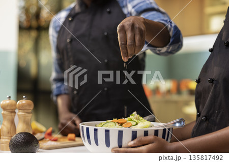 African american man seasoning a salad with salt in a bowl, preparing a healthy lunch at home with organic vegetables. Young vegetarian couple eating fresh produce, wellness and balance. 135817492
