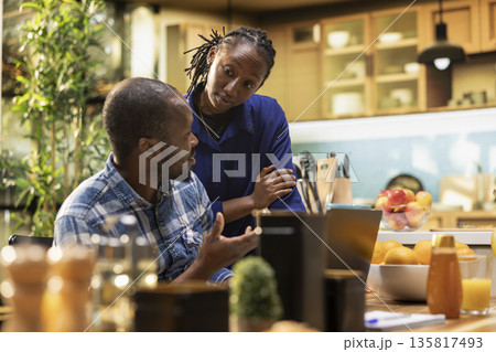 Smiling black man working remotely at the table and chatting with girlfriend, drinking coffee while the boyfriend writes a report on his computer. Teleworking at the breakfast table. 135817493
