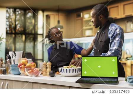 Chroma key screen next to black couple making a salad with fresh ingredients, cutting and mixing vegetables for a vegetarian meal. Young people with aprons bonding in the kitchen. Chroma key screen next to black couple making a salad with fresh ingredients, cutting and mixing vegetables for a vegetarian meal. Young people with aprons bonding in the kitchen. 135817496