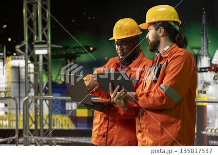 Offshore platform crew members using laptop to analyze extraction efficiency during aurora borealis. Engineers on drilling rig deck configuring vessel management systems under green arctic lights 135817537