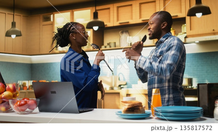 Energetic man and woman singing and laughing in the kitchen, enjoying relaxed morning at home with music. Fun romantic activity shows love, satisfaction and modern lifestyle full of joy. 135817572