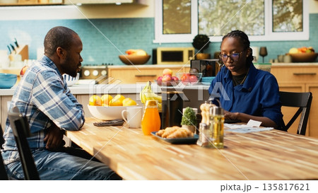 Young couple tracking expenses and doing their household finances, sitting together at kitchen table with receipts, calculator and laptop. Calculating bills, responsible fiscal planning. 135817621