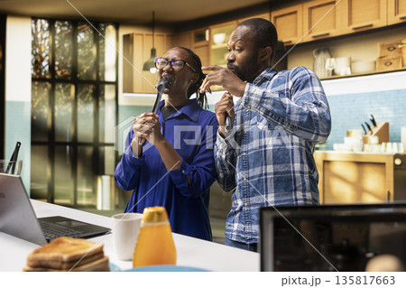 Goofy cheerful people singing together with tools as microphones in the kitchen, black couple showing love and playful energy, fooling around and having fun with music. Positive relations. Goofy cheerful people singing together with tools as microphones in the kitchen, black couple showing love and playful energy, fooling around and having fun with music. Positive relations. 135817663