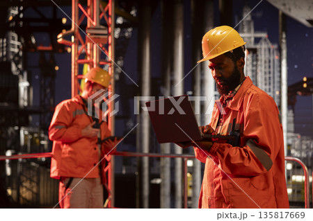 Black offshore oil rig engineer evaluate system performance using apps on laptop, focusing on natural gasoline and oil extraction from industrial pump. Maritime petrol production. Black offshore oil rig engineer evaluate system performance using apps on laptop, focusing on natural gasoline and oil extraction from industrial pump. Maritime petrol production. 135817669