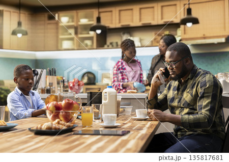 Black young dad on a phone call for work during breakfast meal together, interrupting family bonding time and morning routine to answer important calls. Father neglecting his children. 135817681