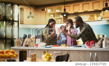 Portrait of black family clapping hands and cheering after finishing homemade pizza in the kitchen. High five and teamwork with laughter shows joy of family cooking and bonding. Camera A. Portrait of black family clapping hands and cheering after finishing homemade pizza in the kitchen. High five and teamwork with laughter shows joy of family cooking and bonding. Camera A. 135817720