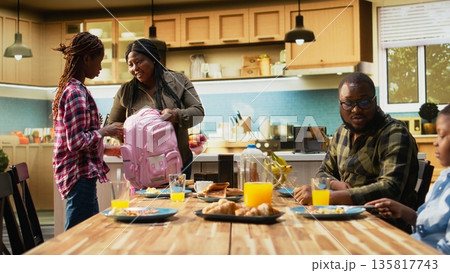 African American mother packing lunch box for her two school kids, preparing their backpack with essentials and snacks for the day. Enjoying rich breakfast with family at home. Camera A. African American mother packing lunch box for her two school kids, preparing their backpack with essentials and snacks for the day. Enjoying rich breakfast with family at home. Camera A. 135817743