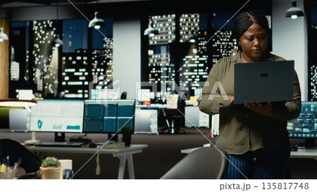 Black specialist reading emails and documenting data from insights for a multinational company, working after hours in a dark office. Focuses on enterprise objectives and development. Camera B. Black specialist reading emails and documenting data from insights for a multinational company, working after hours in a dark office. Focuses on enterprise objectives and development. Camera B. 135817748