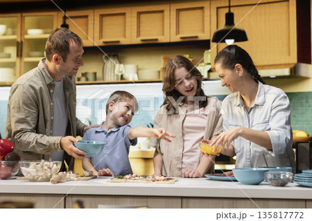 Parents and two children enjoy cooking time in their bright kitchen, working together on a homemade pizza recipe. Family shares laughter making weekend activity a memorable moment. Parents and two children enjoy cooking time in their bright kitchen, working together on a homemade pizza recipe. Family shares laughter making weekend activity a memorable moment. 135817772