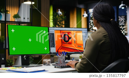Green screen next to african american woman working on game developing, using multiple monitors at the office. Female engineer creating a futuristic spaceship station on software. Camera B. Green screen next to african american woman working on game developing, using multiple monitors at the office. Female engineer creating a futuristic spaceship station on software. Camera B. 135817797