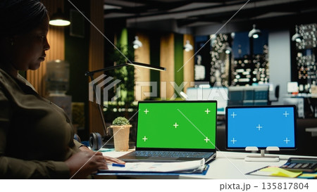 Chroma key on laptop and tablet next to black general manager engaged in budgeting or financial projections at night time. Businesswoman solving important tasks with devices. Camera A. 135817804