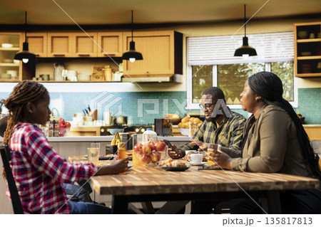 Black preoccupied father being focused on his remote work and neglecting his children at breakfast, ignoring his small kids and wife while eating homemade meal. Distracted distant dad. 135817813