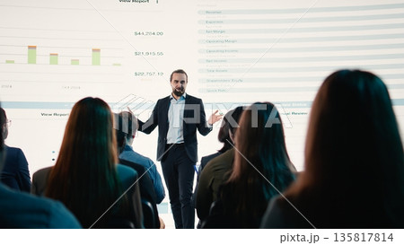 Professional businessman on stage delivering motivational speech during business conference event. Diverse professionals and white collar workers attend international summit in auditorium. 135817814