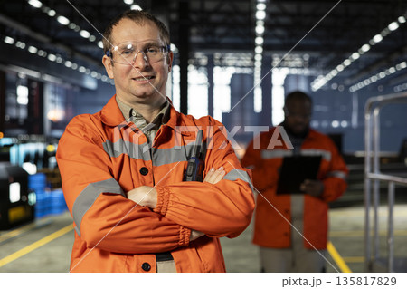 Industrial personnel with safety vest and safety glasses in fabrication workshop, overseeing machinery as sparks rise from metal welding. Manufacturing process and workforce labor. Industrial personnel with safety vest and safety glasses in fabrication workshop, overseeing machinery as sparks rise from metal welding. Manufacturing process and workforce labor. 135817829