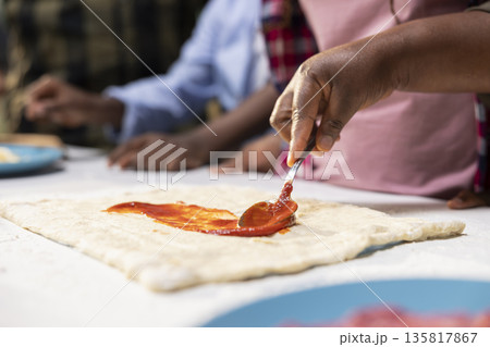 Close up of children spreading tomato sauce on pizza dough at home, making homemade meal with fresh ingredients and produce. Adding dressing as first step, family time in the kitchen. Close up of children spreading tomato sauce on pizza dough at home, making homemade meal with fresh ingredients and produce. Adding dressing as first step, family time in the kitchen. 135817867