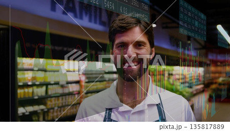 Smiling male grocery clerk wearing apron and showcasing stock charts with tickers in store aisle 135817889