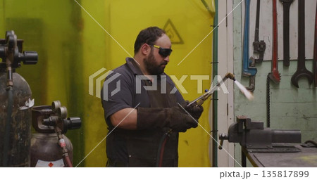 Operating metalworker in leather apron heating vise in workshop, with blowtorch and gas cylinders Operating metalworker in leather apron heating vise in workshop, with blowtorch and gas cylinders 135817899